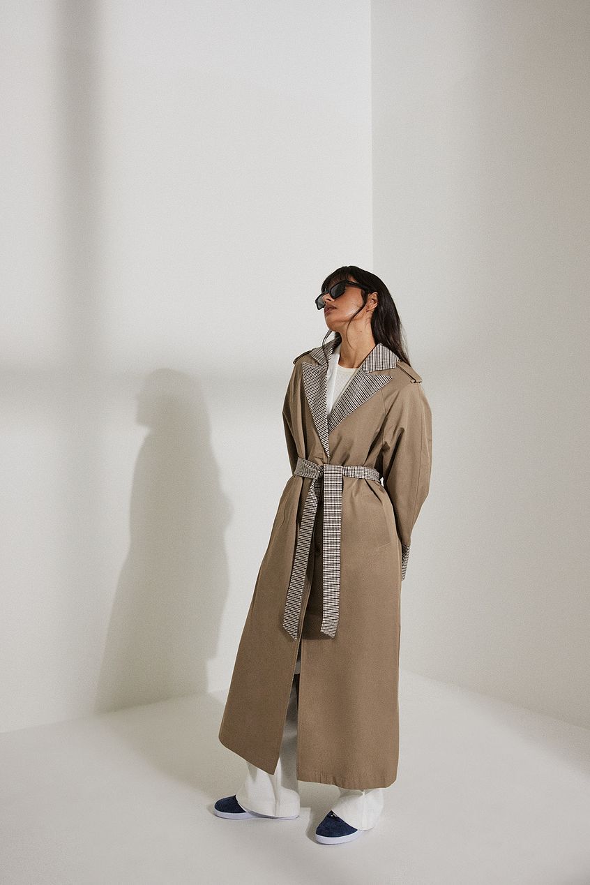 This NA-KD x sanna jornvik trenchcoat is belted and features side slant pockets. It has a button closure and a buckle detail on each shoulder. This trenchcoat features a back slit detail and houndstooth details along the item. This trenchcoat comes in dark beige.