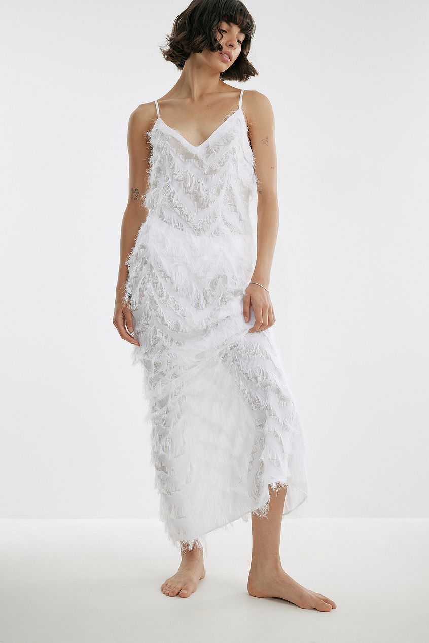NA-KD sheer feathered maxi dress in white. This stunning dress features thin, adjustable shoulder straps, a v neckline, a back zip closure, a back slit detail and a sheer material with a feathered look. This maxi dress comes in white and is also available in black.